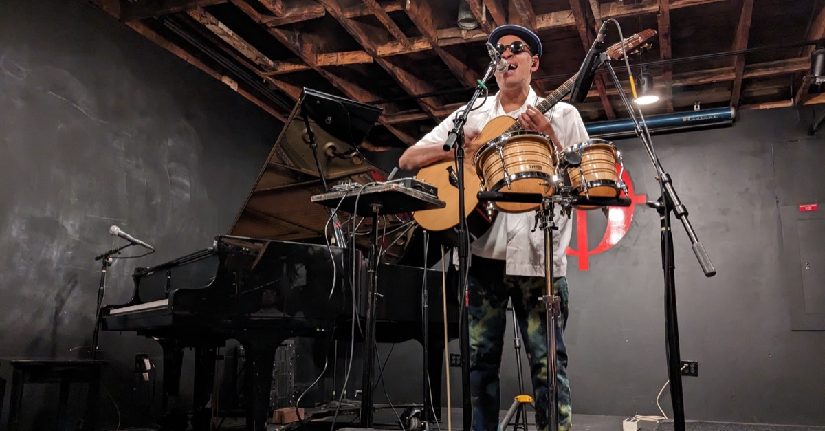 Raul Midón stands on stage playing his guitar, with bongos in front of him and a piano off to one side